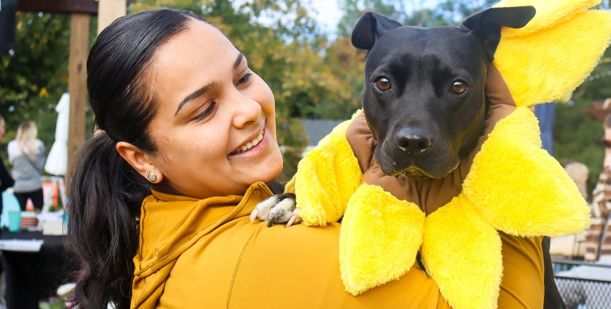 Woman in a yellow costume holding a black dog dressed as a banana.
